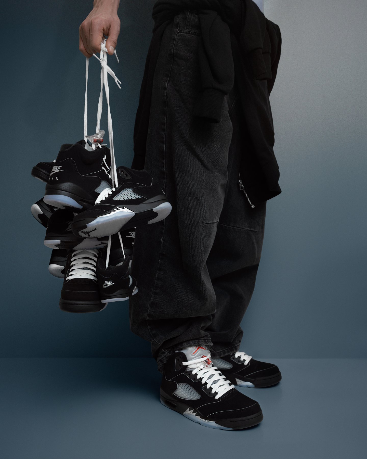 Sneaker shoot: Model holding the black pair by the laces against a blue-grey backdrop